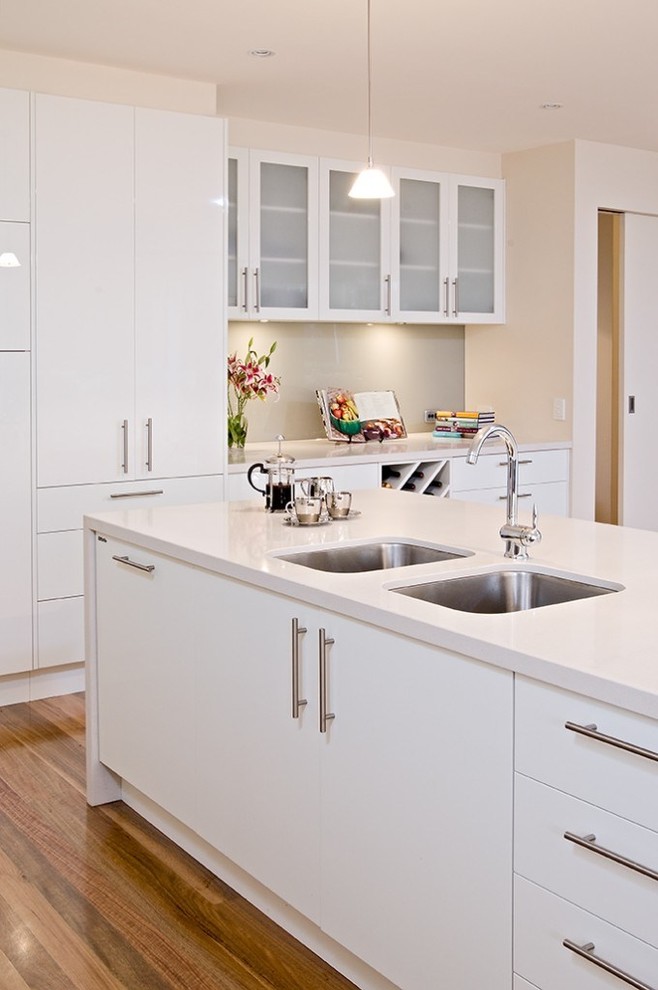 Large modern l-shaped open plan kitchen in Melbourne with an undermount sink, flat-panel cabinets, white cabinets, quartz benchtops, grey splashback, glass sheet splashback, stainless steel appliances, medium hardwood floors and with island.
