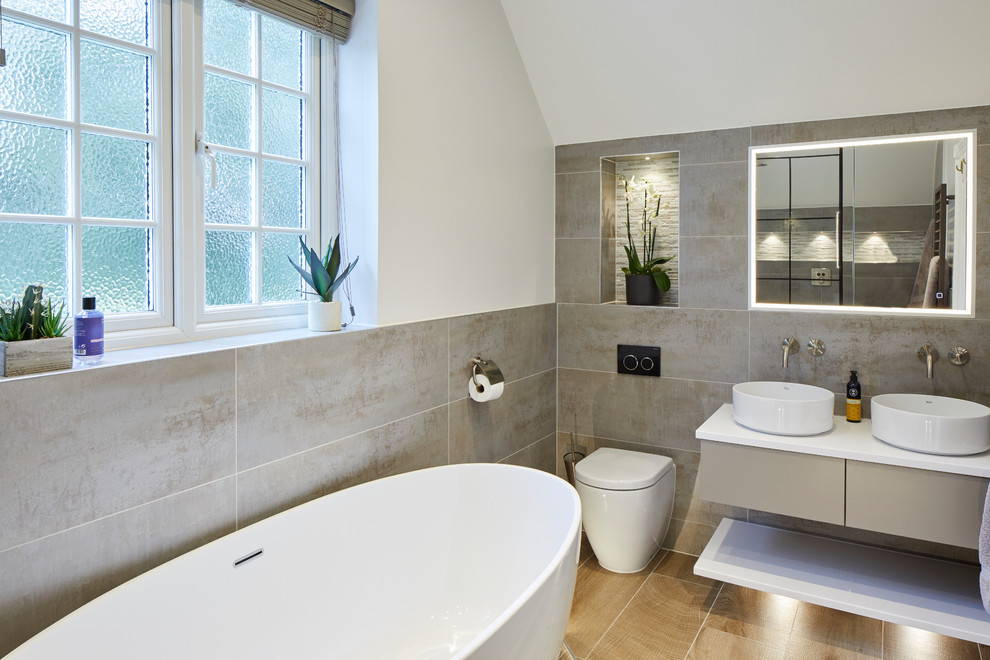 This is an example of a mid-sized industrial master bathroom in Surrey with gray tile, porcelain tile, flat-panel cabinets, grey cabinets, a freestanding tub, white walls, a vessel sink, brown floor and white benchtops.