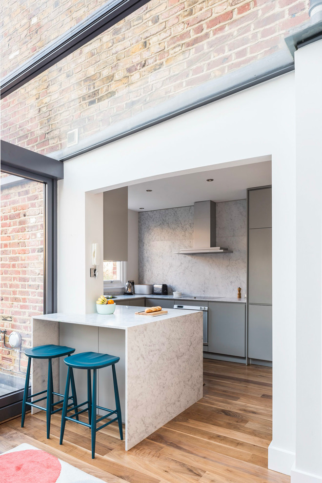 Mid-sized contemporary u-shaped eat-in kitchen in London with an integrated sink, flat-panel cabinets, grey cabinets, marble benchtops, white splashback, marble splashback, stainless steel appliances, light hardwood floors, a peninsula and brown floor.
