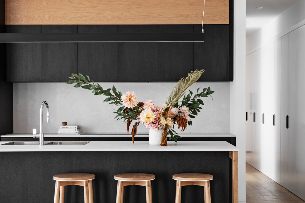 Inspiration for a modern galley kitchen in Wollongong with a drop-in sink, flat-panel cabinets, black cabinets, white splashback, stone slab splashback, stainless steel appliances, medium hardwood floors, with island, brown floor and white benchtop.