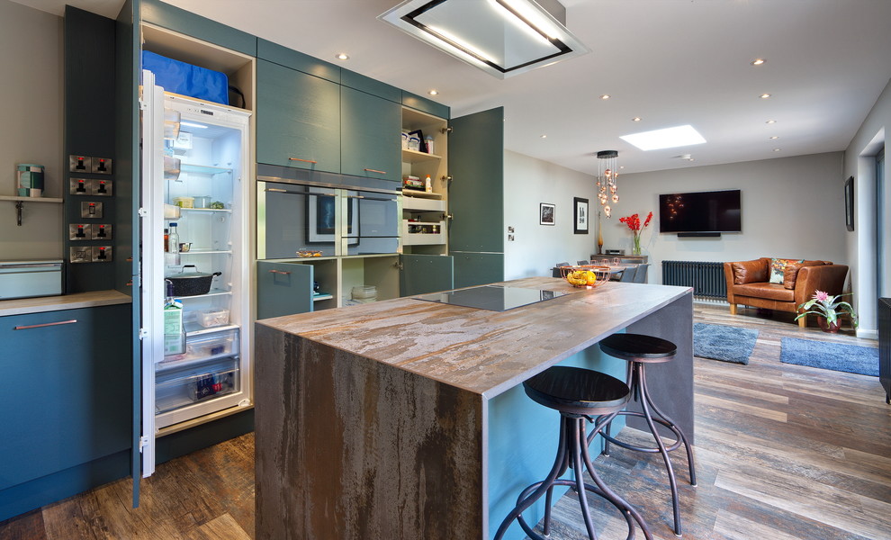 This is an example of a mid-sized contemporary l-shaped open plan kitchen in Other with a drop-in sink, flat-panel cabinets, green cabinets, tile benchtops, grey splashback, timber splashback, stainless steel appliances, laminate floors, with island, brown floor and brown benchtop.