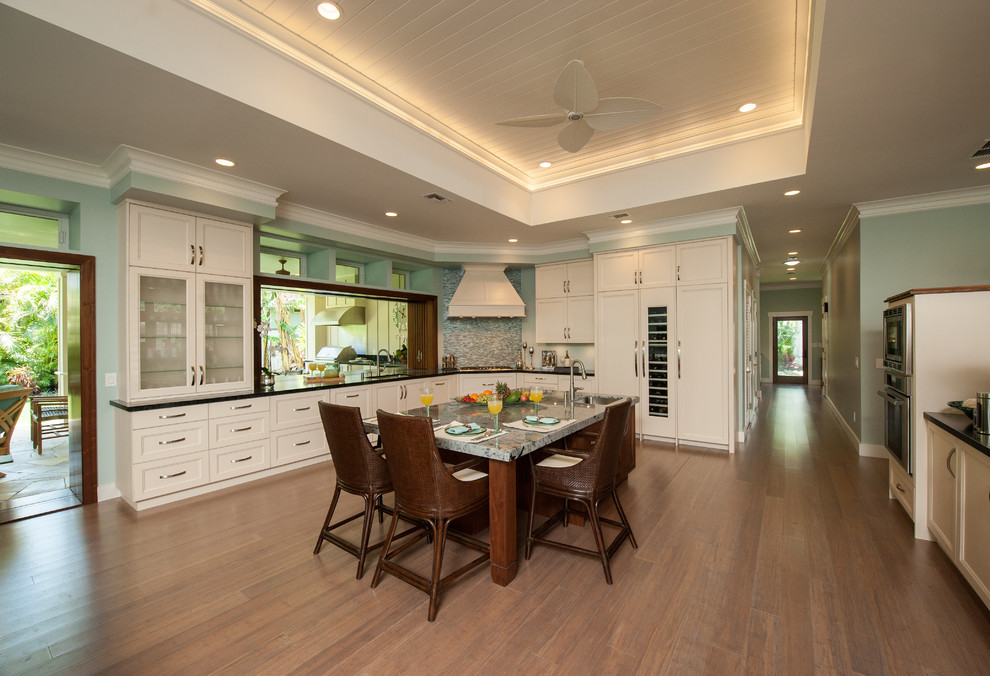 Large transitional l-shaped open plan kitchen in Hawaii with an undermount sink, shaker cabinets, white cabinets, soapstone benchtops, blue splashback, glass sheet splashback, panelled appliances, bamboo floors and with island.