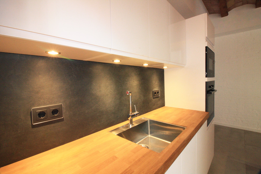 Photo of a mid-sized modern single-wall open plan kitchen in Barcelona with an undermount sink, recessed-panel cabinets, white cabinets, wood benchtops, black splashback, slate splashback, stainless steel appliances, cement tiles, with island and multi-coloured floor.