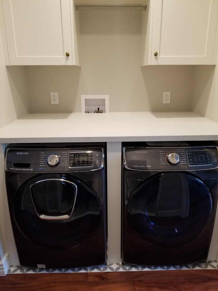 Photo of a small modern single-wall laundry cupboard in Los Angeles with shaker cabinets, white cabinets, solid surface benchtops, beige walls, medium hardwood floors, a side-by-side washer and dryer, brown floor and white benchtop.