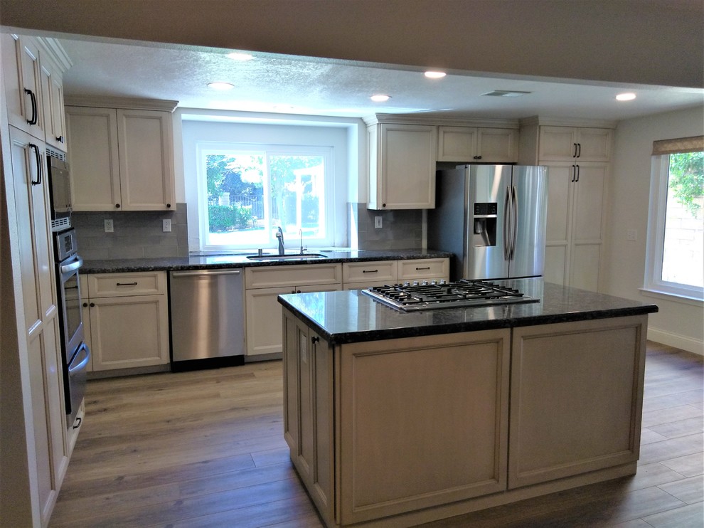 This is an example of a mid-sized transitional galley open plan kitchen in Los Angeles with an undermount sink, recessed-panel cabinets, beige cabinets, granite benchtops, grey splashback, ceramic splashback, stainless steel appliances, vinyl floors, with island, brown floor and black benchtop.