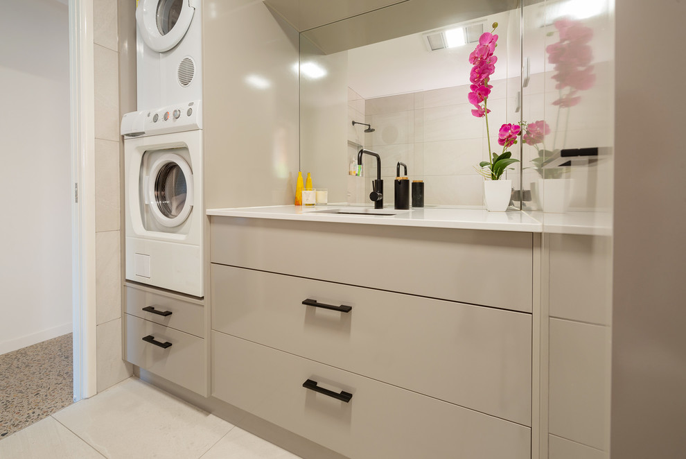 Photo of a mid-sized contemporary single-wall utility room in Brisbane with quartz benchtops, porcelain floors, a stacked washer and dryer, beige floor, an undermount sink, flat-panel cabinets, beige cabinets, beige walls and white benchtop.