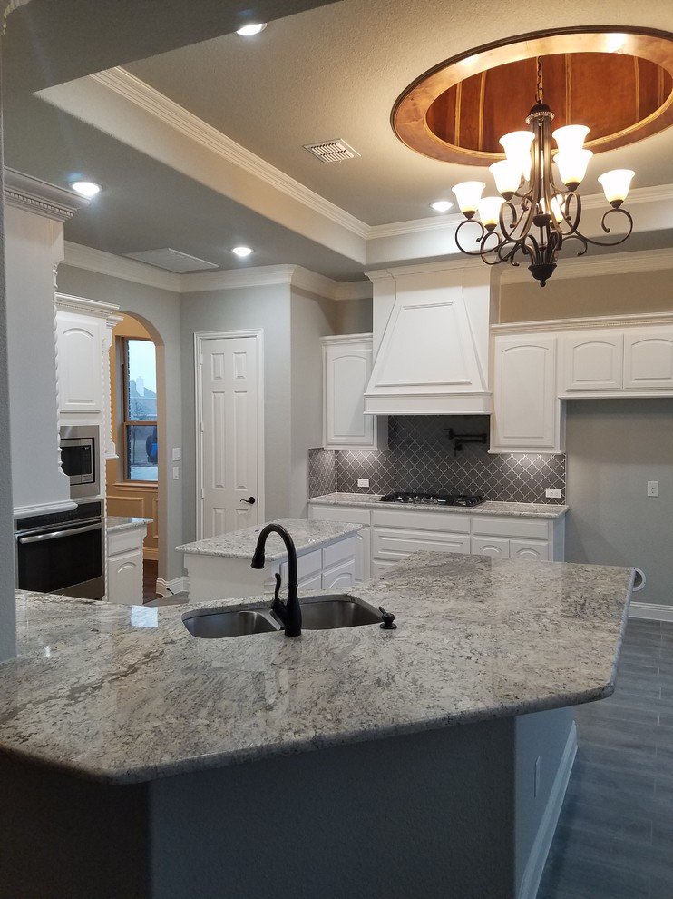 This is an example of a large traditional l-shaped open plan kitchen in Dallas with a double-bowl sink, raised-panel cabinets, white cabinets, granite benchtops, grey splashback, porcelain splashback, stainless steel appliances, dark hardwood floors, with island, brown floor and grey benchtop.