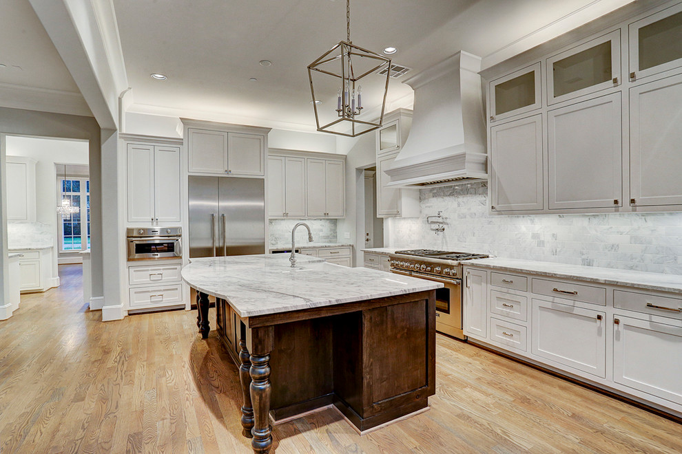 This is an example of a large arts and crafts l-shaped open plan kitchen in Houston with a farmhouse sink, louvered cabinets, grey cabinets, granite benchtops, grey splashback, stone tile splashback, stainless steel appliances, light hardwood floors, with island and brown floor.