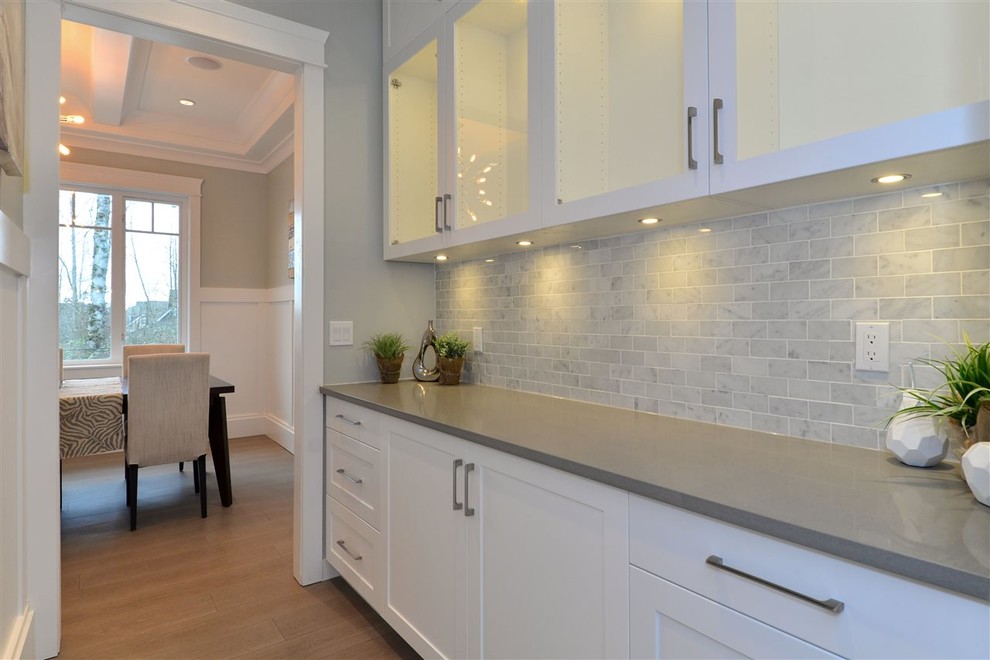 Photo of a mid-sized contemporary single-wall home bar in Vancouver with no sink, glass-front cabinets, white cabinets, quartz benchtops, grey splashback, marble splashback, light hardwood floors, beige floor and grey benchtop.