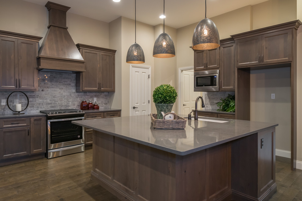 Large mountain style galley medium tone wood floor and brown floor open concept kitchen photo with a single-bowl sink, shaker cabinets, brown cabinets, quartz countertops, gray backsplash, stone tile backsplash, stainless steel appliances and an island