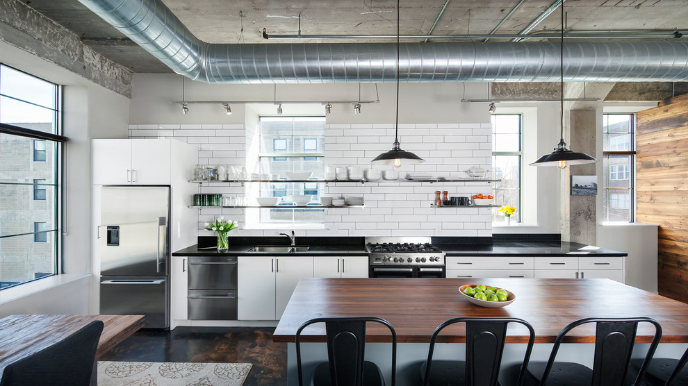 Inspiration for a large industrial single-wall eat-in kitchen in Minneapolis with flat-panel cabinets, white cabinets, white splashback, subway tile splashback, concrete floors, with island, a double-bowl sink, granite benchtops and stainless steel appliances.