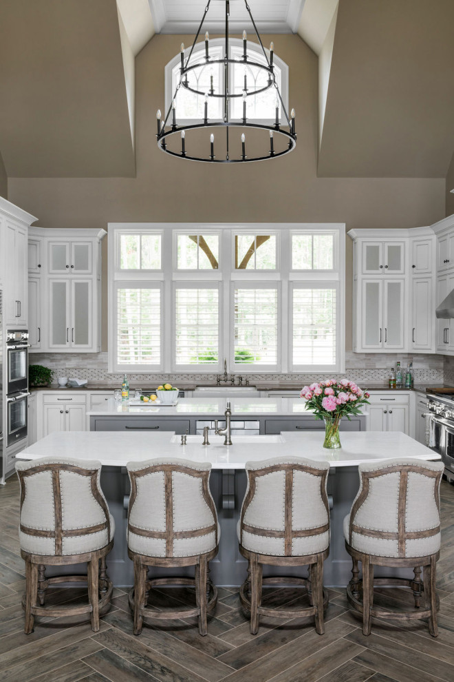 This is an example of an expansive u-shaped open plan kitchen in Atlanta with a farmhouse sink, recessed-panel cabinets, white cabinets, quartz benchtops, grey splashback, marble splashback, stainless steel appliances, porcelain floors, multiple islands, brown floor, white benchtop and vaulted.