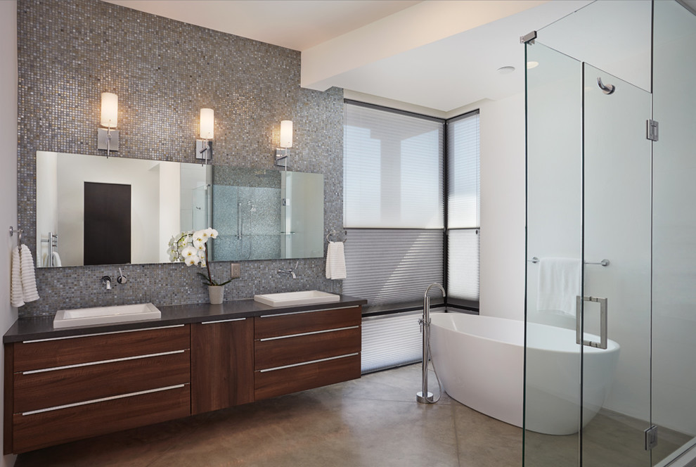 Photo of a modern master bathroom in Phoenix with flat-panel cabinets, brown cabinets, a freestanding tub, gray tile, glass tile, white walls, concrete floors, a wall-mount sink and engineered quartz benchtops.