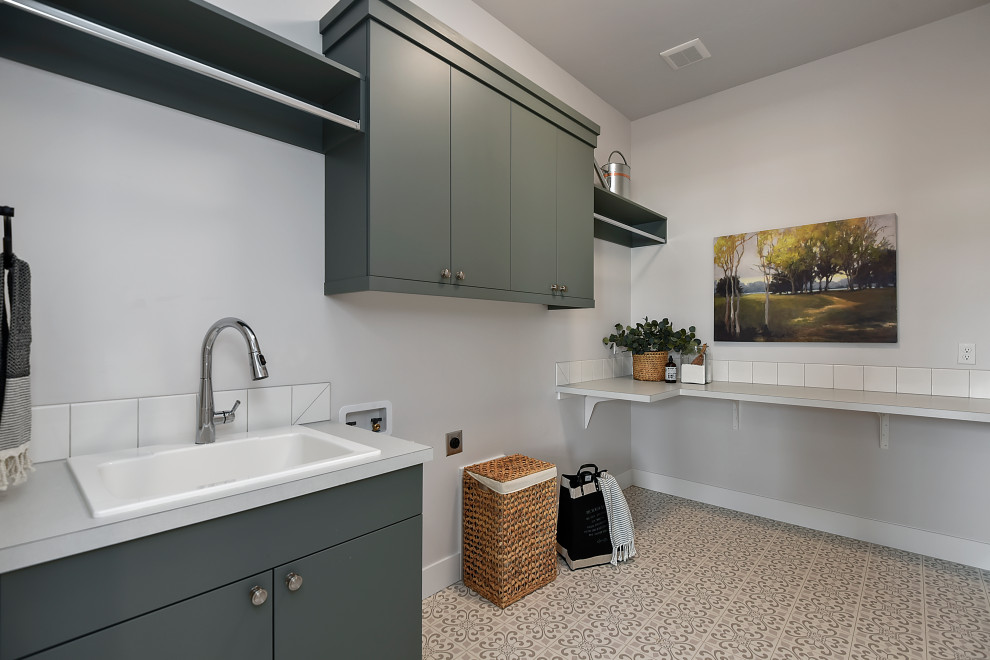 Inspiration for a mid-sized arts and crafts l-shaped utility room in Boise with a drop-in sink, flat-panel cabinets, green cabinets, laminate benchtops, white splashback, porcelain splashback, beige walls, vinyl floors, a side-by-side washer and dryer, multi-coloured floor and grey benchtop.