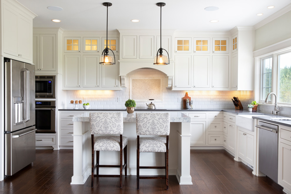 Inspiration for a large arts and crafts u-shaped eat-in kitchen in Milwaukee with shaker cabinets, white cabinets, quartz benchtops, white splashback, ceramic splashback, stainless steel appliances, with island, grey benchtop, a farmhouse sink, dark hardwood floors and brown floor.