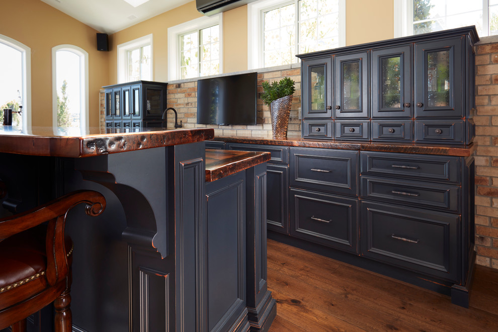 This is an example of a mid-sized country galley seated home bar in Toronto with an undermount sink, raised-panel cabinets, black cabinets, wood benchtops, brown splashback, stone tile splashback and medium hardwood floors.