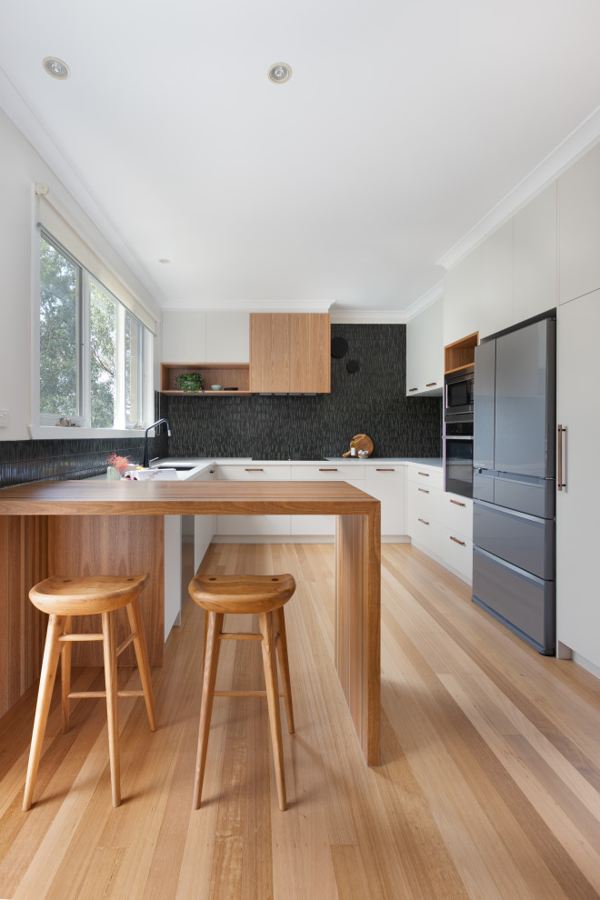 Photo of a contemporary u-shaped kitchen in Melbourne with an undermount sink, flat-panel cabinets, grey cabinets, black splashback, mosaic tile splashback, stainless steel appliances, light hardwood floors, a peninsula, beige floor and white benchtop.