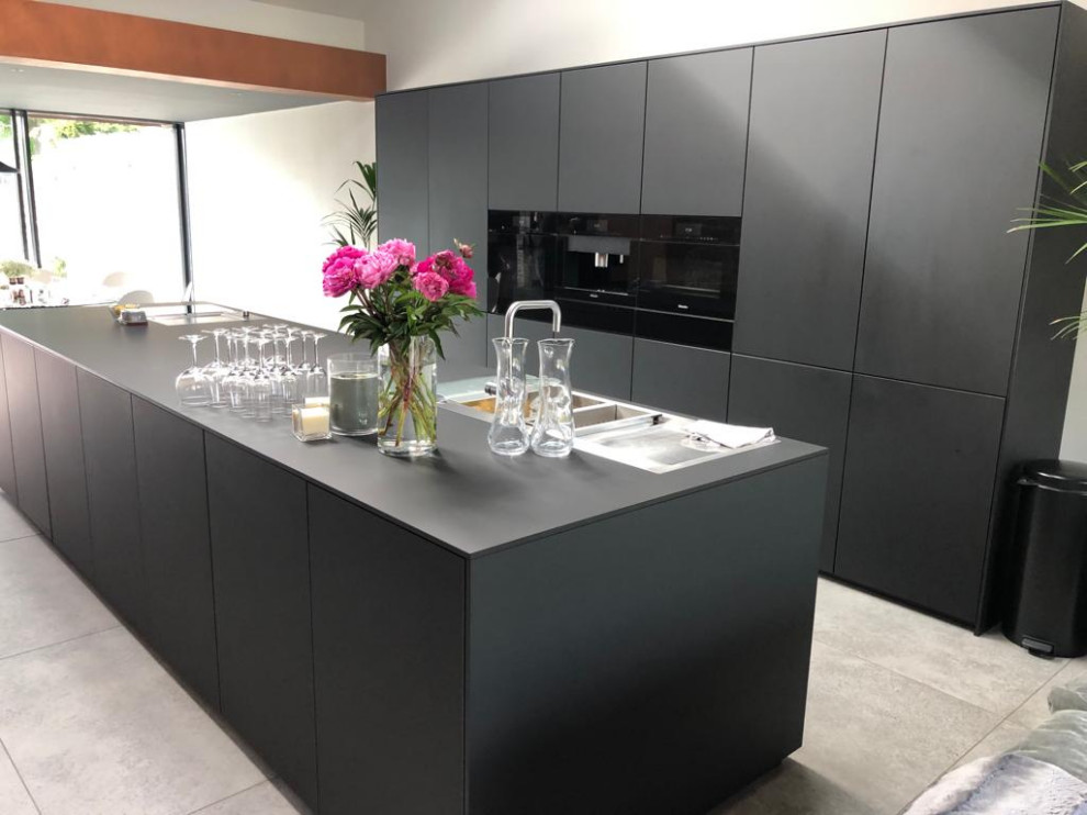 Inspiration for a large contemporary single-wall open plan kitchen in Cardiff with a double-bowl sink, flat-panel cabinets, black cabinets, solid surface benchtops, black appliances, porcelain floors, with island, beige floor and black benchtop.