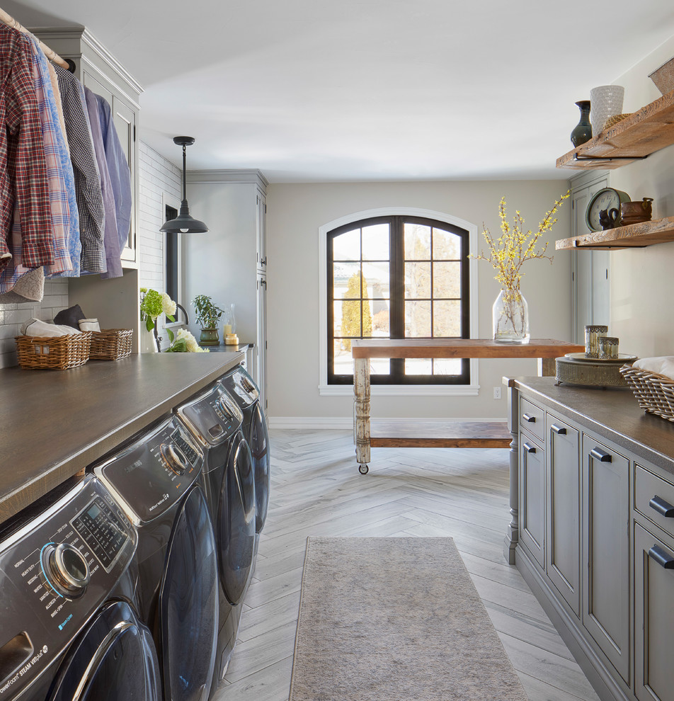 Design ideas for a large country single-wall dedicated laundry room in Denver with a farmhouse sink, recessed-panel cabinets, grey cabinets, soapstone benchtops, white walls, porcelain floors, a side-by-side washer and dryer, grey floor and black benchtop.