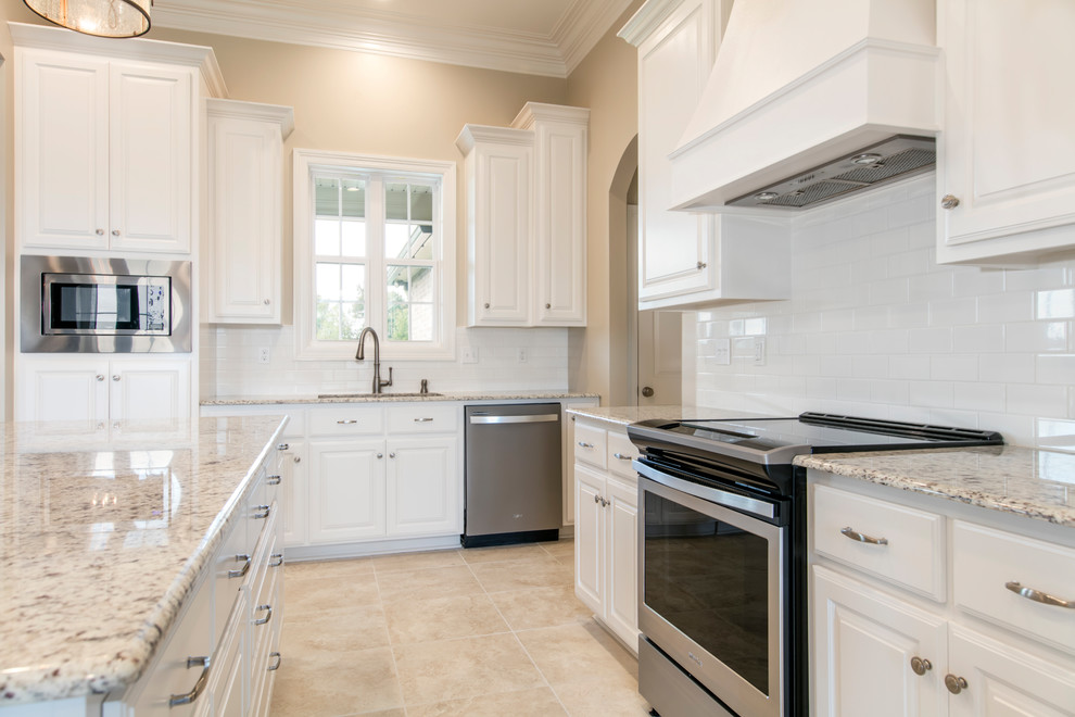 Photo of a mid-sized transitional l-shaped eat-in kitchen in New Orleans with a double-bowl sink, raised-panel cabinets, white cabinets, granite benchtops, white splashback, subway tile splashback, stainless steel appliances, ceramic floors, with island, beige floor and white benchtop.