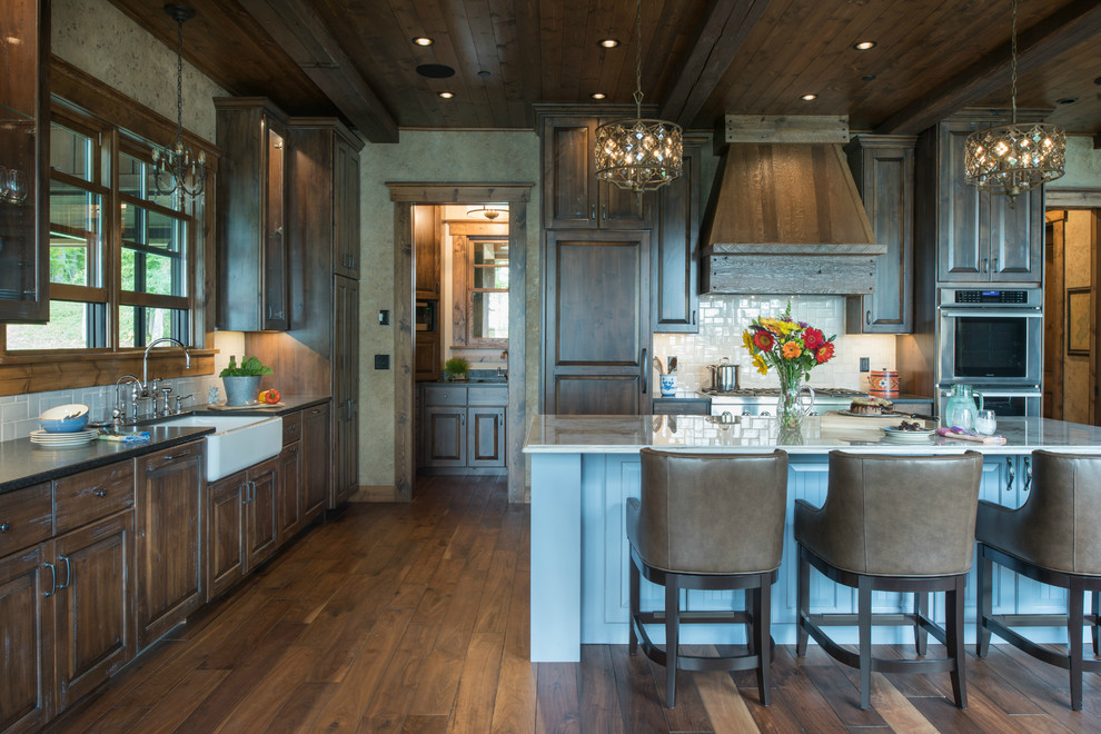 This is an example of a large country l-shaped eat-in kitchen in Minneapolis with a farmhouse sink, raised-panel cabinets, dark wood cabinets, granite benchtops, white splashback, stone tile splashback, panelled appliances, medium hardwood floors, with island and brown floor.