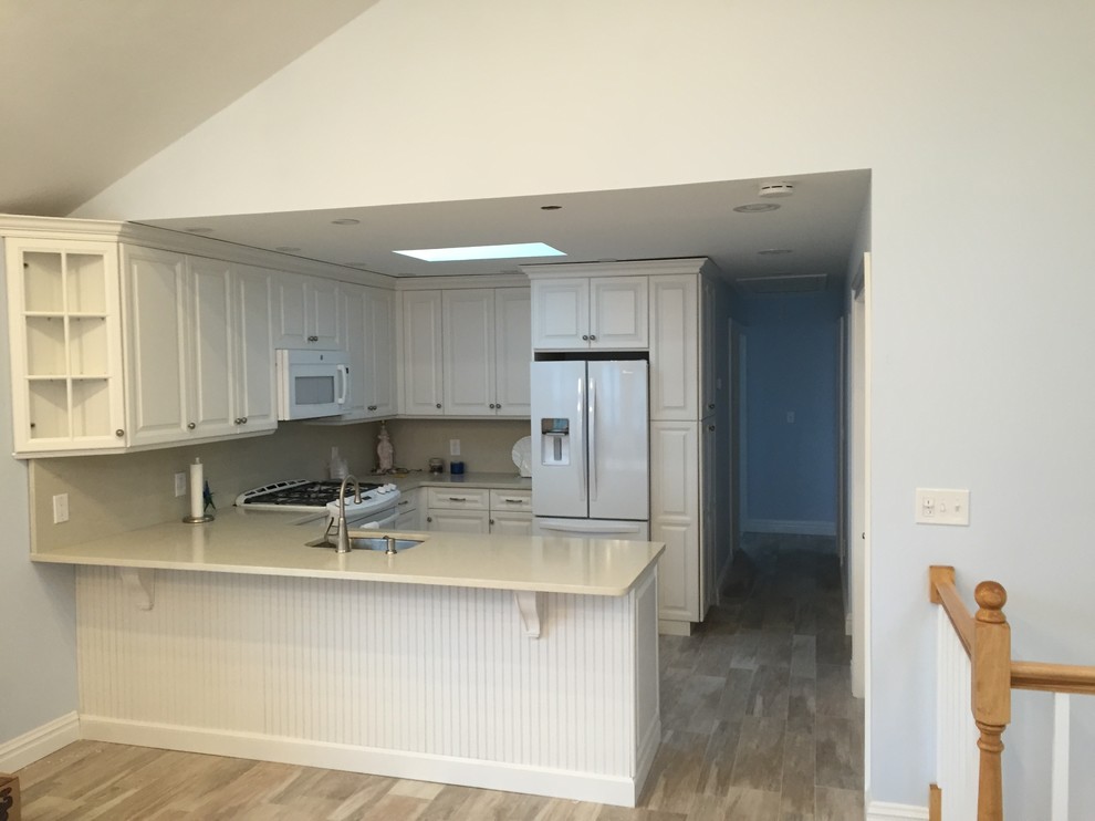 This is an example of a mid-sized beach style u-shaped open plan kitchen in New York with an undermount sink, louvered cabinets, white cabinets, quartz benchtops, beige splashback, white appliances, ceramic floors and a peninsula.