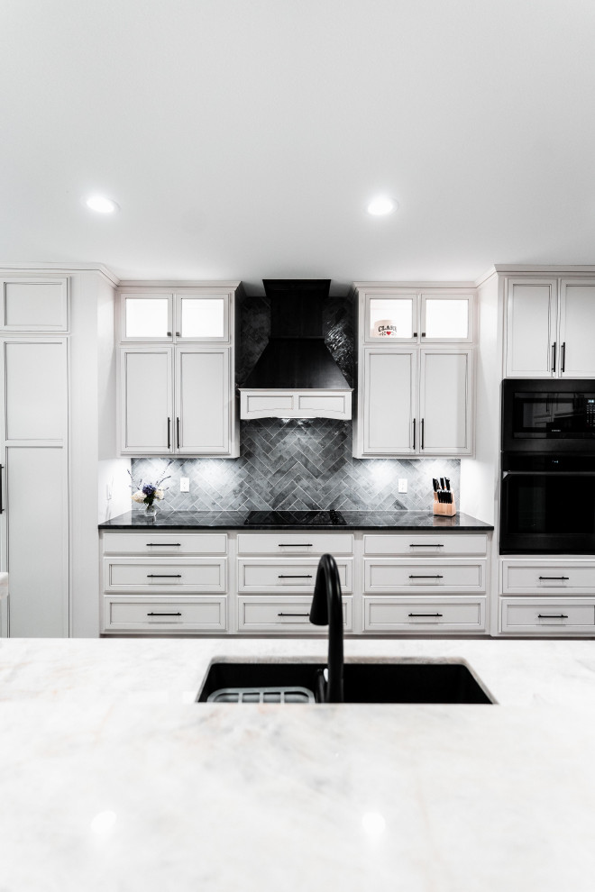 Example of a huge transitional l-shaped laminate floor, gray floor and tray ceiling open concept kitchen design with an undermount sink, flat-panel cabinets, white cabinets, quartz countertops, gray backsplash, ceramic backsplash, black appliances and an island