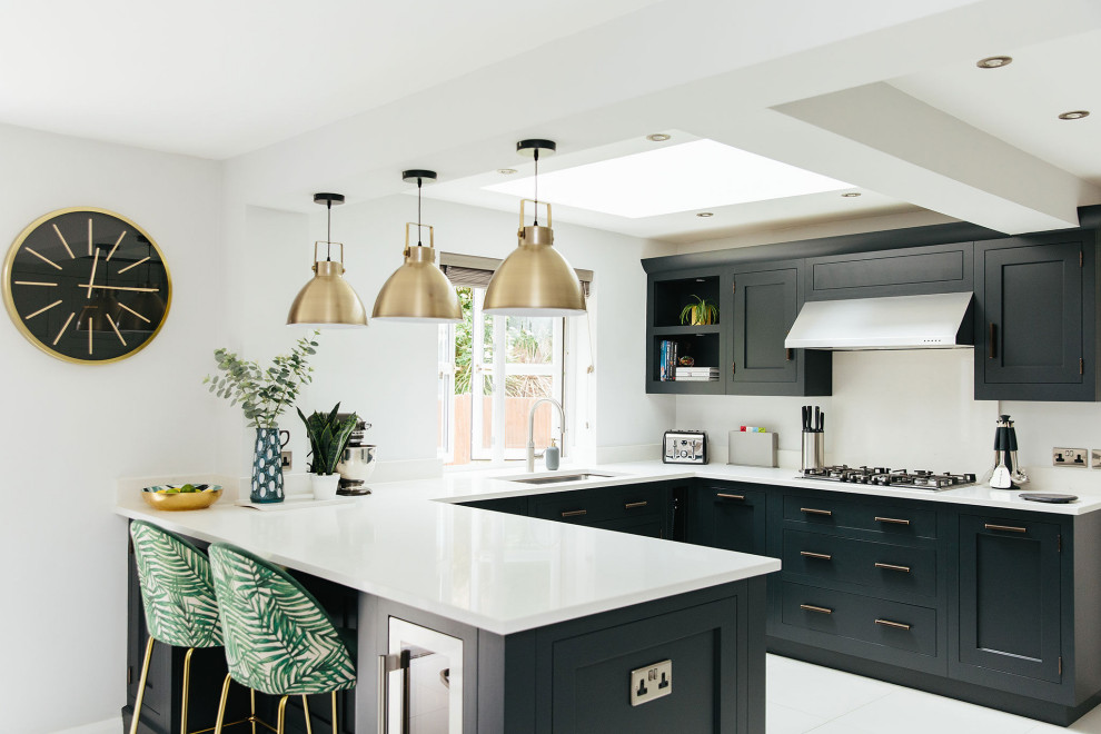 This is an example of a large transitional u-shaped eat-in kitchen in Hertfordshire with a drop-in sink, black cabinets, white splashback, stainless steel appliances, a peninsula and white benchtop.