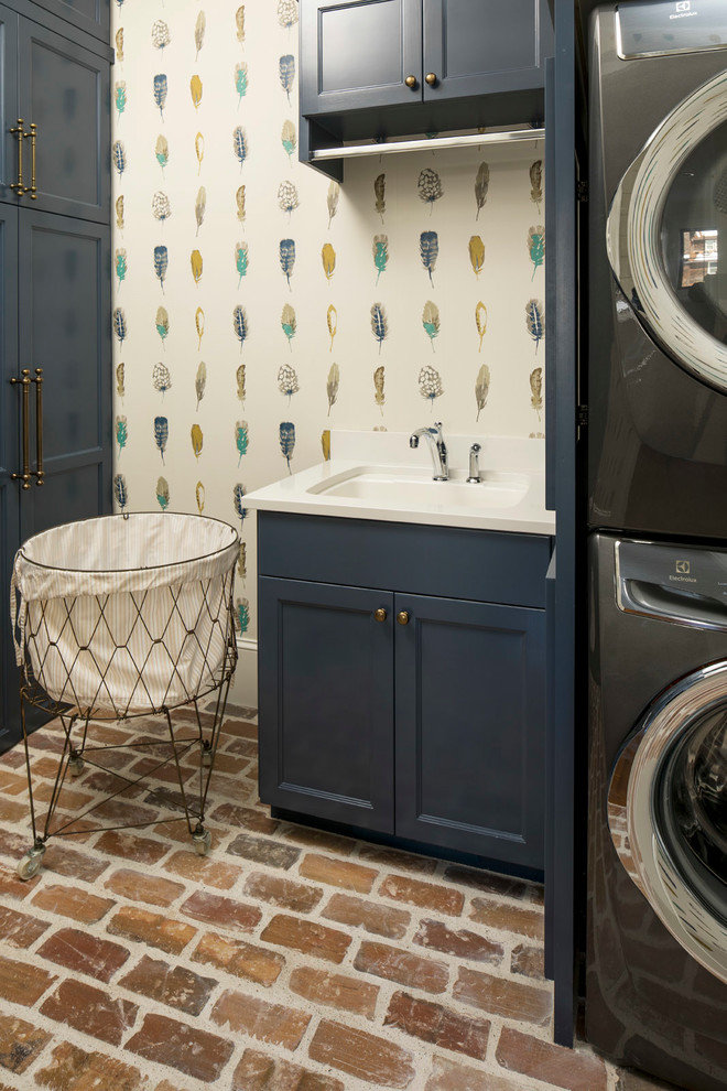 Photo of a small transitional l-shaped dedicated laundry room in Minneapolis with recessed-panel cabinets, blue cabinets, quartz benchtops, multi-coloured walls, brick floors, a stacked washer and dryer and an undermount sink.