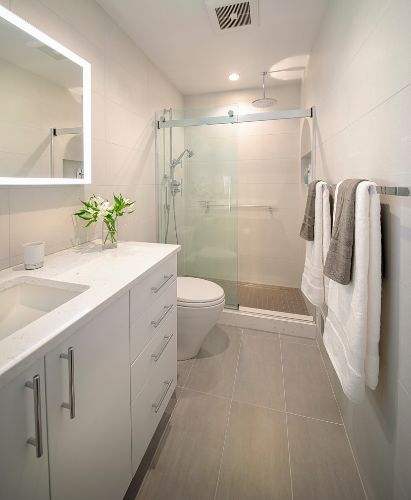 This is an example of a mid-sized modern 3/4 bathroom in DC Metro with flat-panel cabinets, white cabinets, an alcove shower, a one-piece toilet, beige tile, porcelain tile, beige walls, laminate floors, an undermount sink, quartzite benchtops, beige floor, a sliding shower screen and white benchtops.
