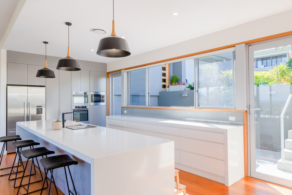 Inspiration for a beach style galley kitchen pantry in Other with an undermount sink, white cabinets, grey splashback, glass sheet splashback, black appliances, light hardwood floors, with island, brown floor and white benchtop.