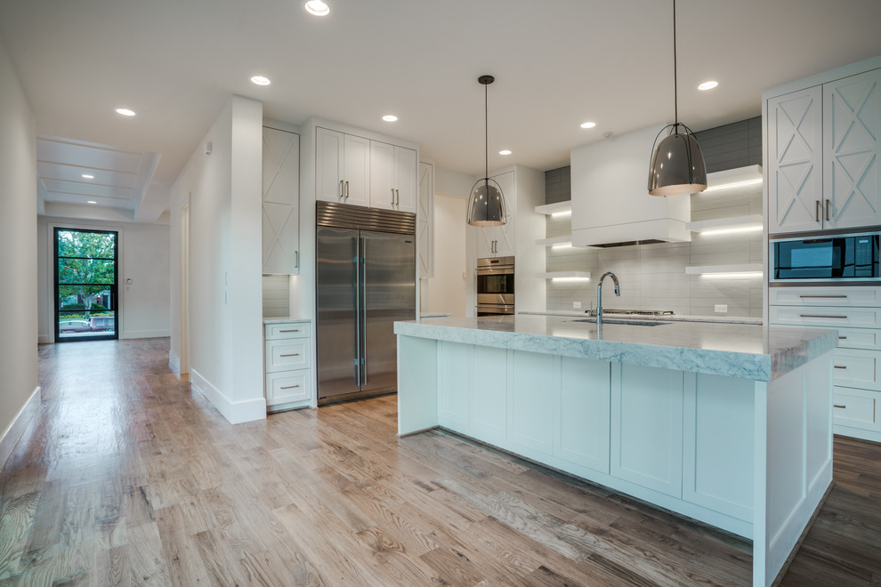 Inspiration for a large transitional single-wall kitchen in Dallas with an undermount sink, shaker cabinets, white cabinets, marble benchtops, white splashback, light hardwood floors, with island and stainless steel appliances.