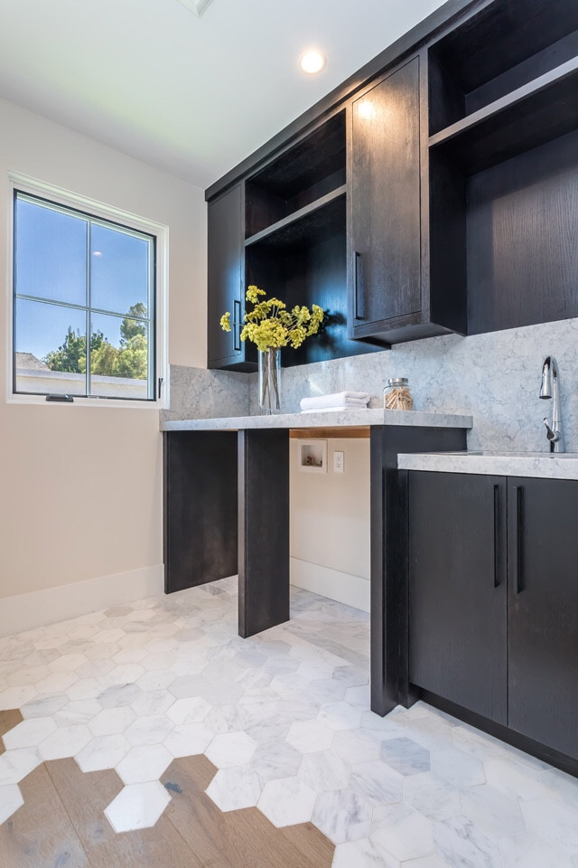 Inspiration for a mid-sized country l-shaped dedicated laundry room with an undermount sink, flat-panel cabinets, black cabinets, marble benchtops, beige walls, a side-by-side washer and dryer, grey benchtop, marble floors and grey floor.