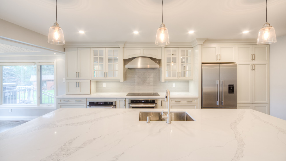 Inspiration for a mid-sized transitional galley light wood floor and beige floor open concept kitchen remodel in Toronto with a double-bowl sink, recessed-panel cabinets, white cabinets, solid surface countertops, white backsplash, porcelain backsplash, stainless steel appliances, an island and white countertops