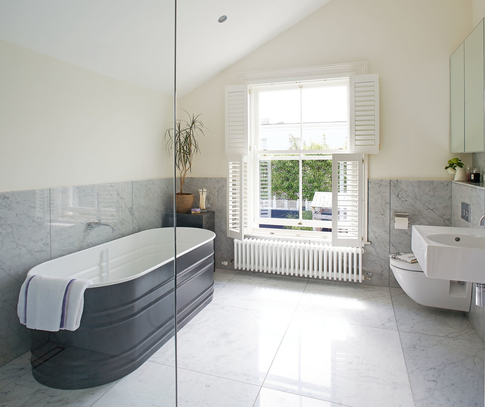 This is an example of a traditional bathroom in Dublin with a freestanding tub, a wall-mount toilet, gray tile, white walls and a wall-mount sink.