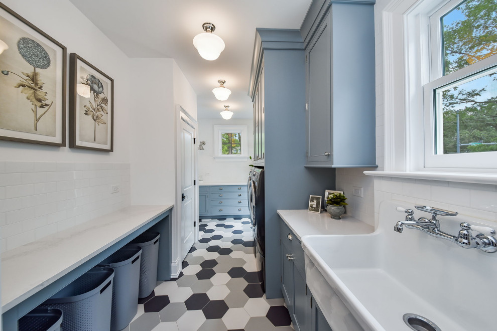 This is an example of a large transitional dedicated laundry room in Chicago with a farmhouse sink, flat-panel cabinets, blue cabinets, quartz benchtops, white splashback, subway tile splashback, white walls, ceramic floors, a side-by-side washer and dryer, blue floor and white benchtop.