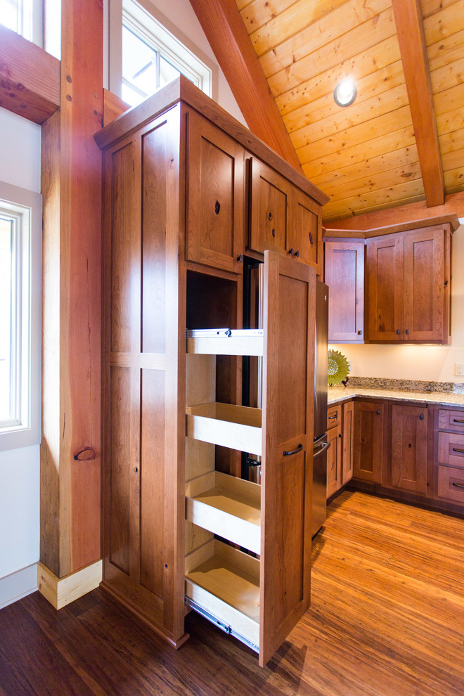 Example of a mountain style l-shaped medium tone wood floor kitchen design with a farmhouse sink, shaker cabinets, medium tone wood cabinets, granite countertops, stainless steel appliances and an island