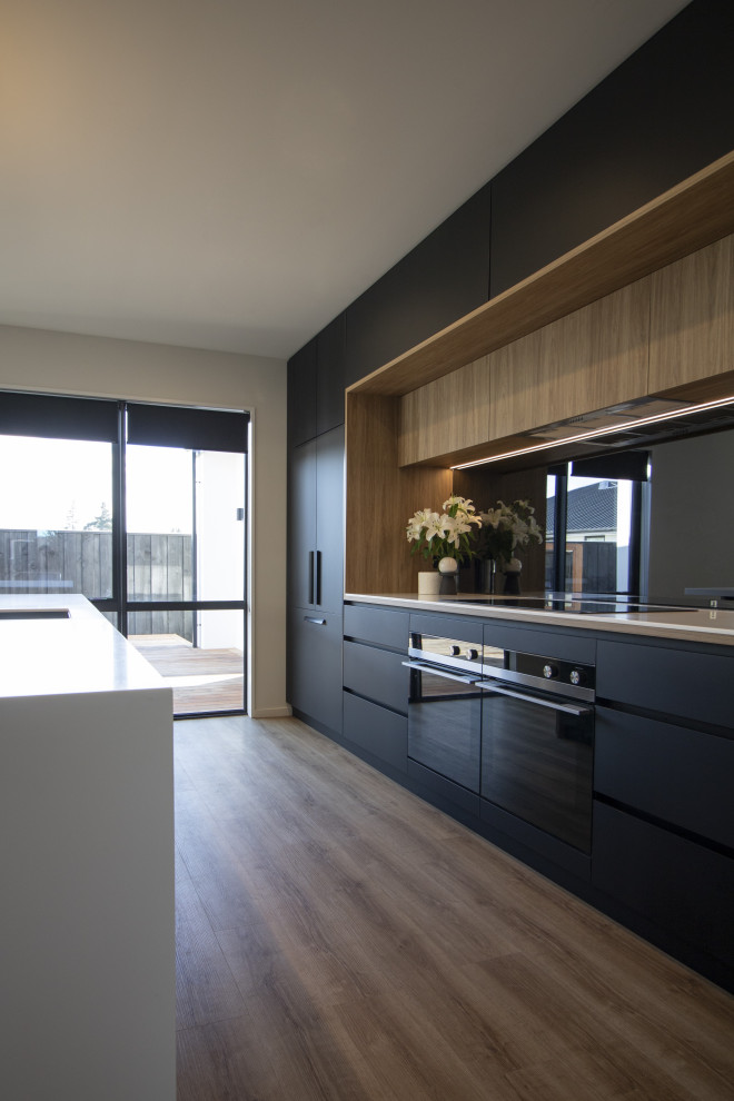 Eat-in kitchen - large modern galley medium tone wood floor and brown floor eat-in kitchen idea in Christchurch with a single-bowl sink, black cabinets, solid surface countertops, metallic backsplash, mirror backsplash, black appliances, an island and white countertops