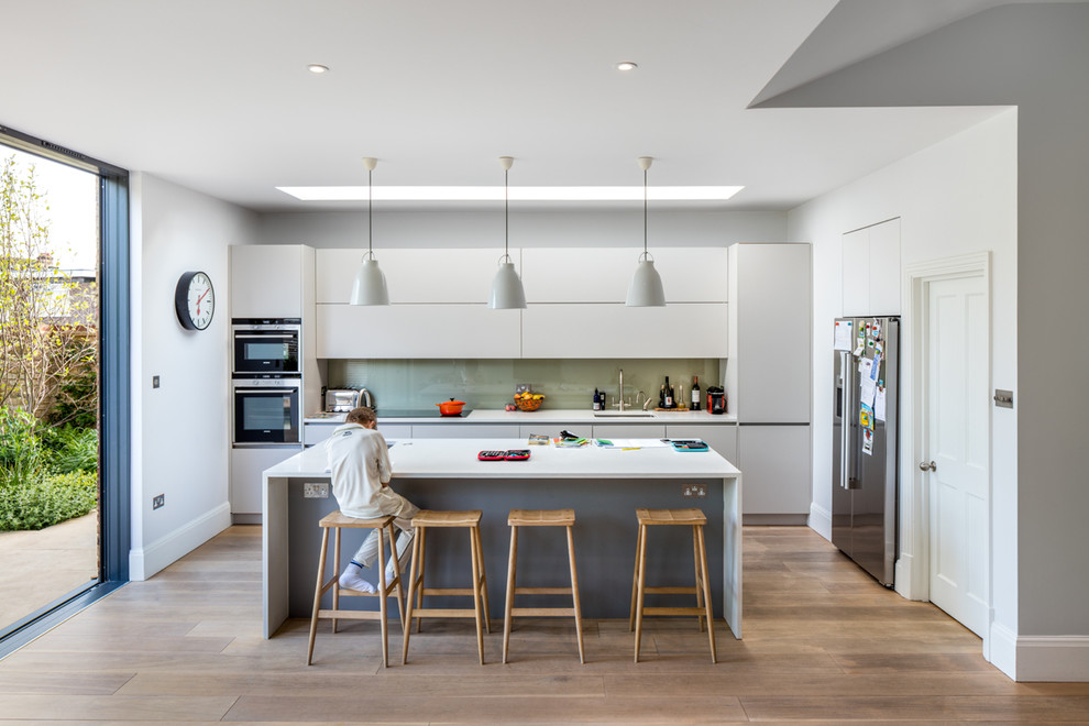 Photo of a large contemporary single-wall eat-in kitchen in London with an integrated sink, flat-panel cabinets, white cabinets, solid surface benchtops, green splashback, glass sheet splashback, panelled appliances, with island, light hardwood floors and brown floor.
