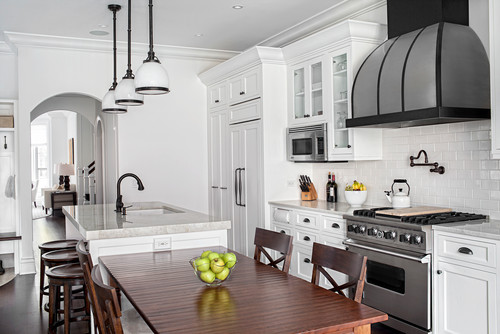 Transitional white kitchen with shaker-style Kitchen Cabinet Mississauga, subway tile backsplash, large black range hood, quartz countertops, and an island beside a wood dining table.