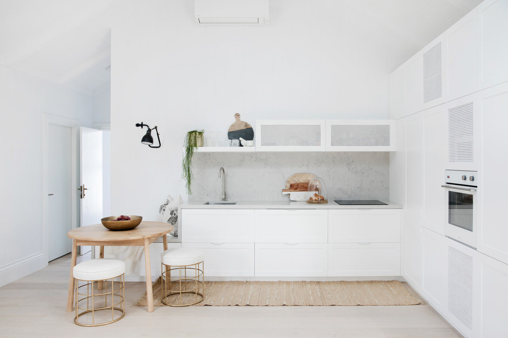 Design ideas for a beach style l-shaped open plan kitchen in Sydney with a single-bowl sink, shaker cabinets, white cabinets, white appliances, no island, grey splashback, light hardwood floors, beige floor and grey benchtop.