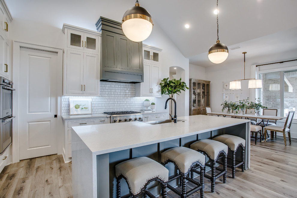 Example of a mid-sized transitional l-shaped medium tone wood floor and brown floor open concept kitchen design in Austin with a farmhouse sink, shaker cabinets, white cabinets, marble countertops, white backsplash, stainless steel appliances, an island and white countertops