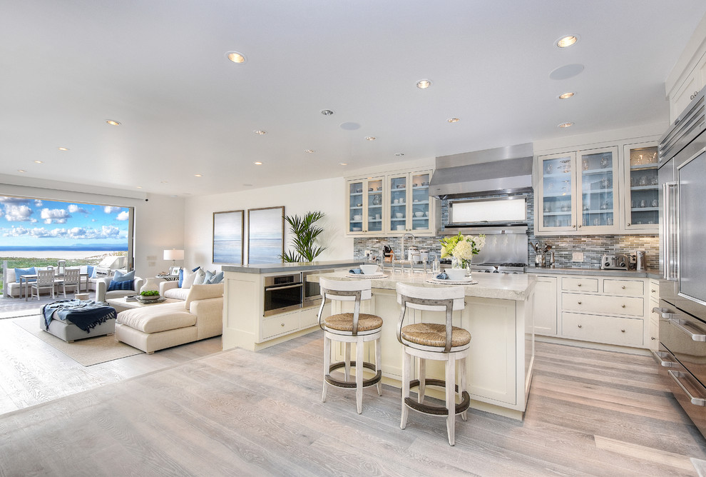 This is an example of a mid-sized beach style u-shaped open plan kitchen in Orange County with glass-front cabinets, white cabinets, with island, stainless steel appliances, a farmhouse sink, grey splashback, matchstick tile splashback, grey floor and light hardwood floors.