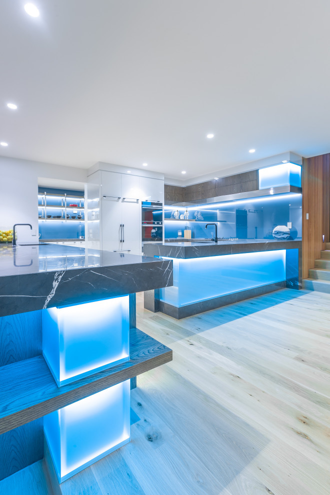 Huge trendy l-shaped light wood floor and brown floor open concept kitchen photo in Auckland with an undermount sink, flat-panel cabinets, white cabinets, granite countertops, blue backsplash, glass sheet backsplash, stainless steel appliances, two islands and gray countertops
