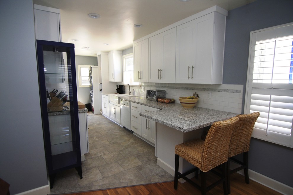 Photo of a mid-sized modern galley eat-in kitchen in Los Angeles with a farmhouse sink, shaker cabinets, white cabinets, granite benchtops, white splashback, ceramic splashback, stainless steel appliances, travertine floors and no island.