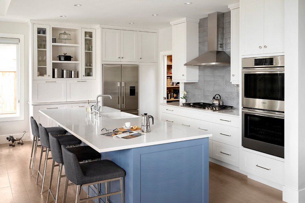 Eat-in kitchen - mid-sized transitional galley medium tone wood floor and brown floor eat-in kitchen idea in Vancouver with a double-bowl sink, shaker cabinets, white cabinets, gray backsplash, stainless steel appliances, an island, quartz countertops and ceramic backsplash