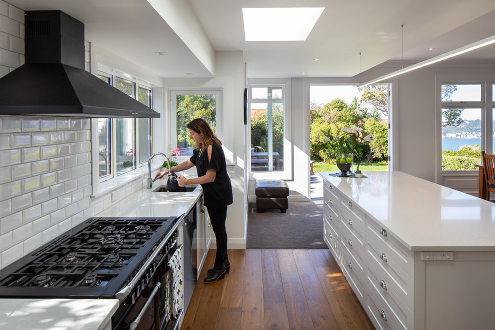Inspiration for a galley open plan kitchen in Wellington with an undermount sink, shaker cabinets, white cabinets, solid surface benchtops, white splashback, subway tile splashback, black appliances, medium hardwood floors, with island, brown floor and white benchtop.