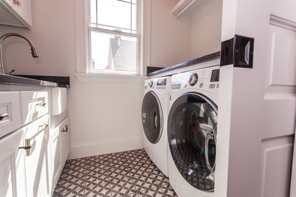 Photo of a small transitional galley dedicated laundry room in Cincinnati with a farmhouse sink, raised-panel cabinets, white cabinets, granite benchtops, white walls, porcelain floors, a side-by-side washer and dryer, multi-coloured floor and black benchtop.