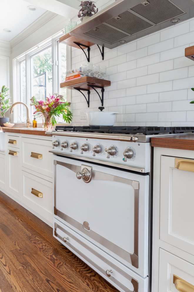 Design ideas for a large eclectic galley eat-in kitchen in Chicago with a farmhouse sink, recessed-panel cabinets, white cabinets, wood benchtops, white splashback, subway tile splashback, stainless steel appliances, medium hardwood floors, with island, brown floor and brown benchtop.