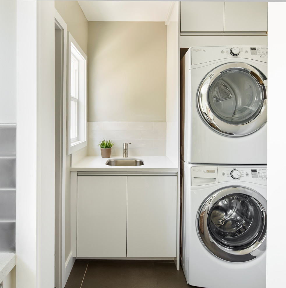This is an example of a contemporary laundry room in Los Angeles with flat-panel cabinets, white cabinets, beige walls, a stacked washer and dryer, brown floor and white benchtop.